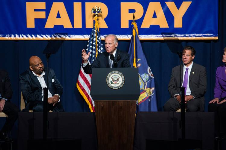 President Biden speaking at a podium in front of a sign that says "Fair Pay"